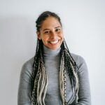 Smiling ethnic female with long braids in turtleneck and jeans crossing hands standing against white background